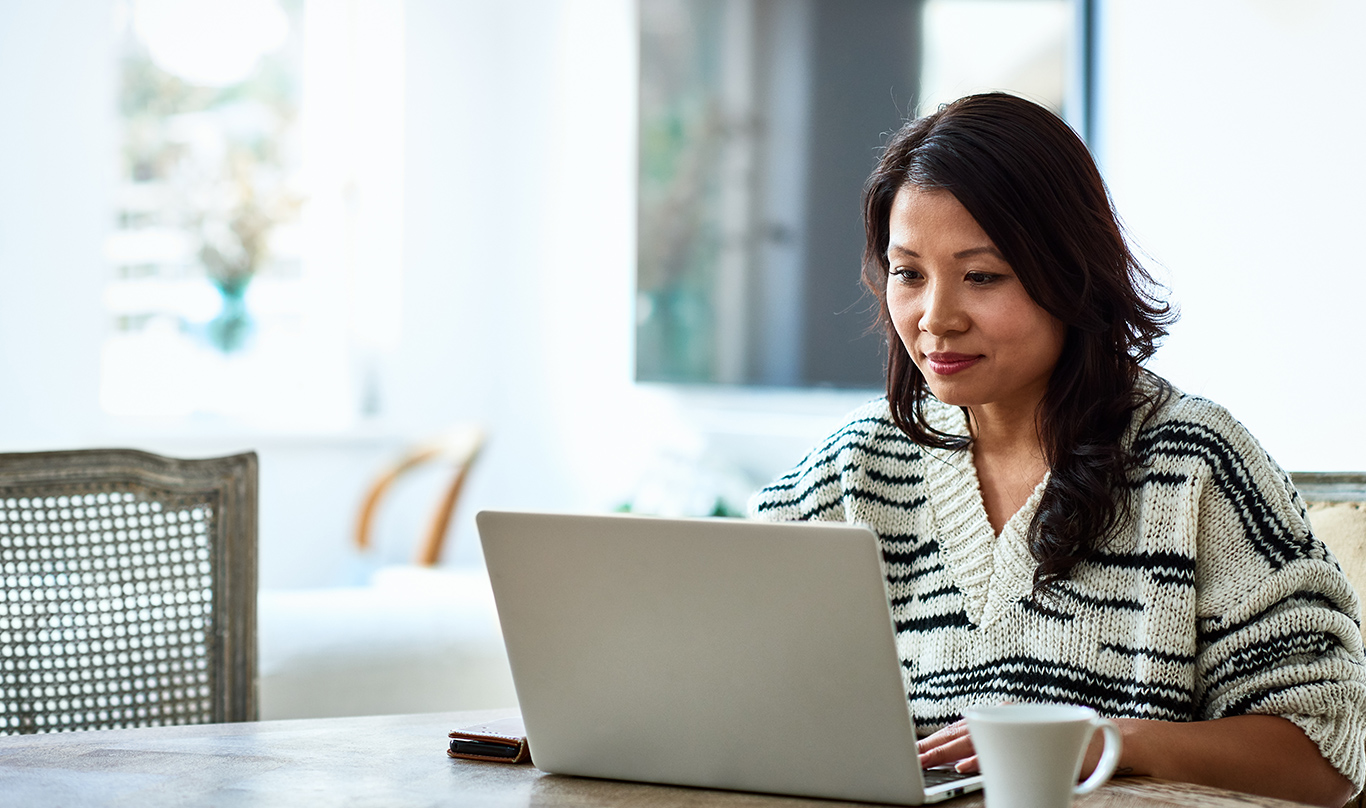 A woman in a sweater works on a laptop at her kitchen table.