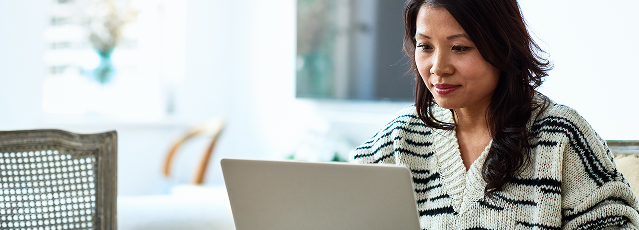 A woman in a sweater works on a laptop at her kitchen table.