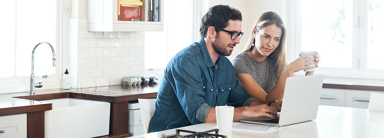 Couple sitting at counter in kitchen with mugs, looking at a laptop computer