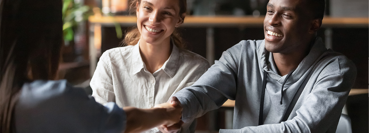 A happy man reaches out to shake a person's hand across a desk. A happy woman looks on.