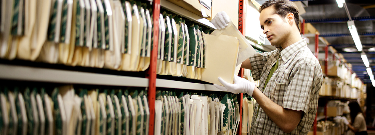 A man wears gloves while reviewing an old document in an archive.