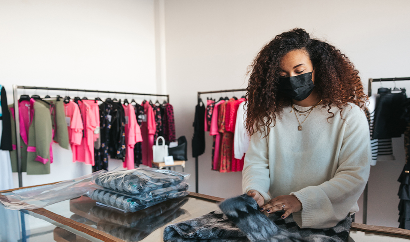 An associate at a retail clothing shop folds a weather while wearing a mask.