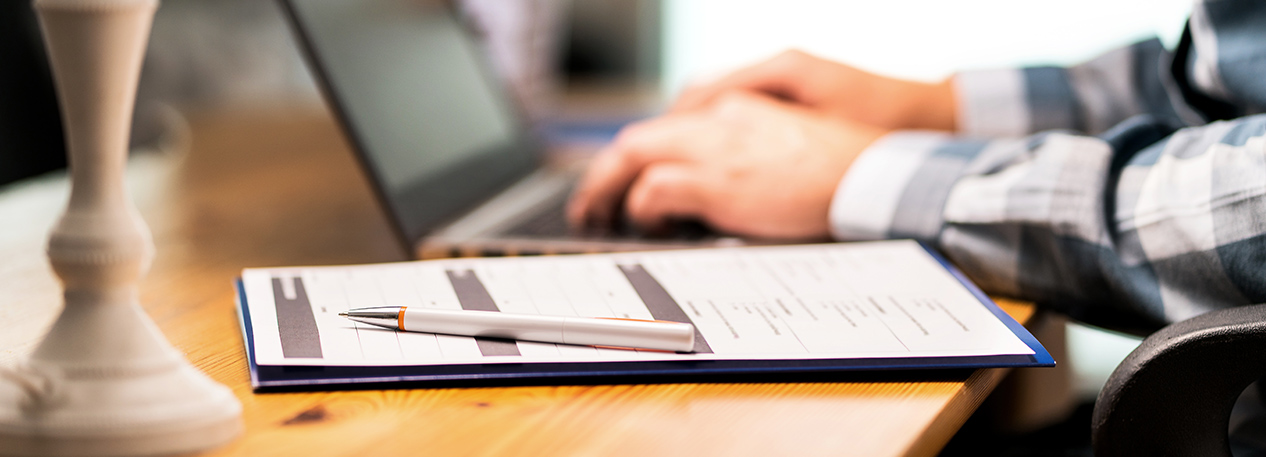 Documents and a pen on a desk, with person working on a laptop computer in the background