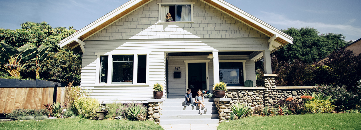 Two children sit on the front porch of a white house while another looks down from a window above.