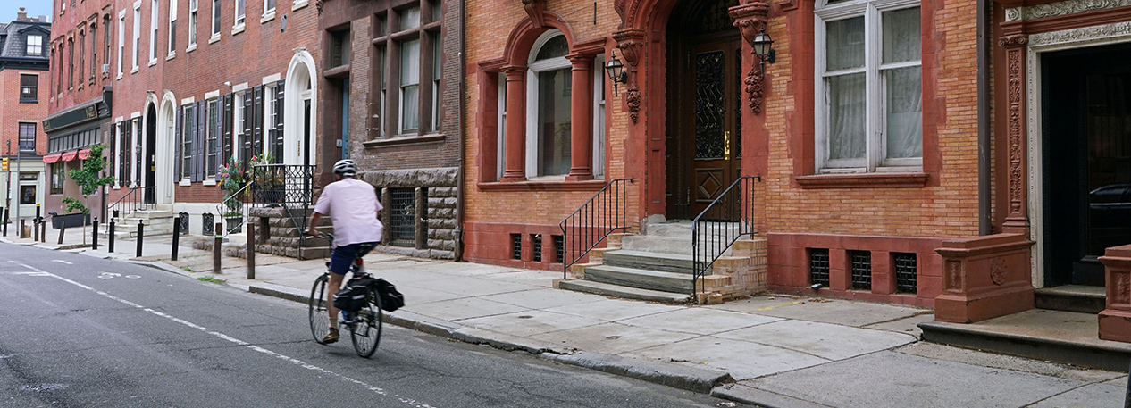 A man rides a bike down a city street along a block of brick townhouses.