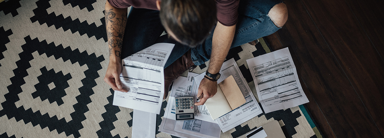 Man sitting cross-legged on the floor, using a calculator, surrounded by printed forms