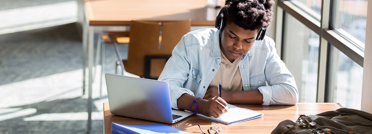 Student with headphones sitting at a small table with a laptop computer, writing in a notebook