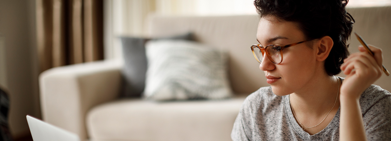 Woman sitting on the floor in front of a couch, holding a pen and looking at a laptop computer