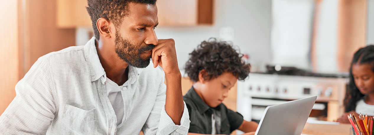 A dad looks at his open laptop with a worried look while his son plays in the background.
