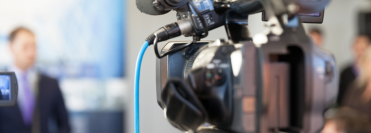 Back of a broadcast video camera, with a reporter in a suit in the distance
