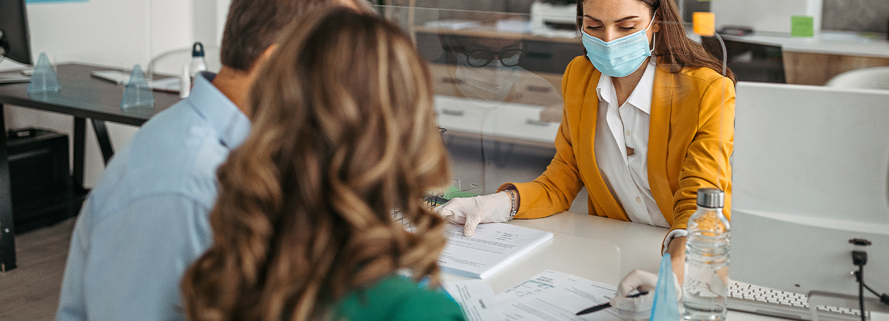 Man and woman reviewing forms at a desk with a woman who is wearing a mask.