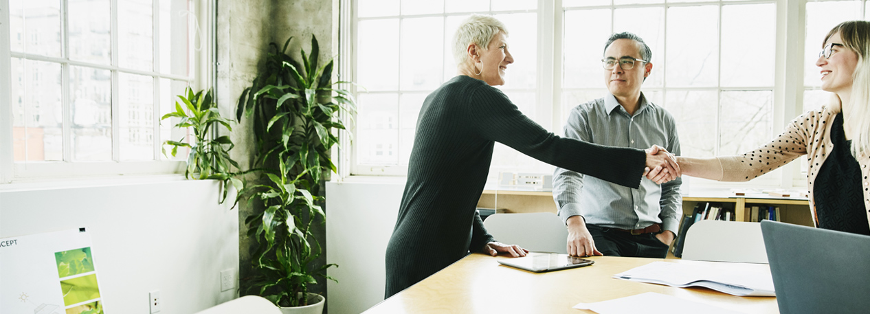 Standing at a conference table, two women shake hands while an intermediary observes.