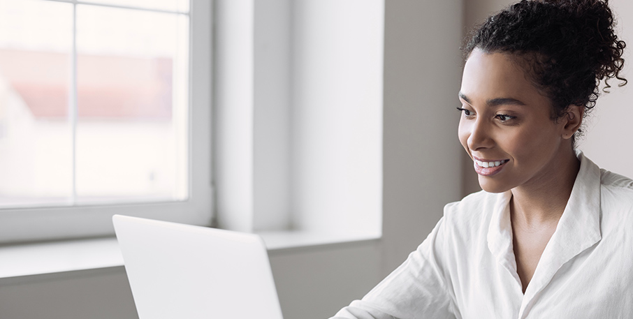 A business woman dressed in white works on a laptop in front of a window.