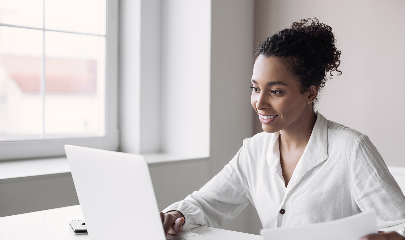 A business woman dressed in white works on a laptop in front of a window.