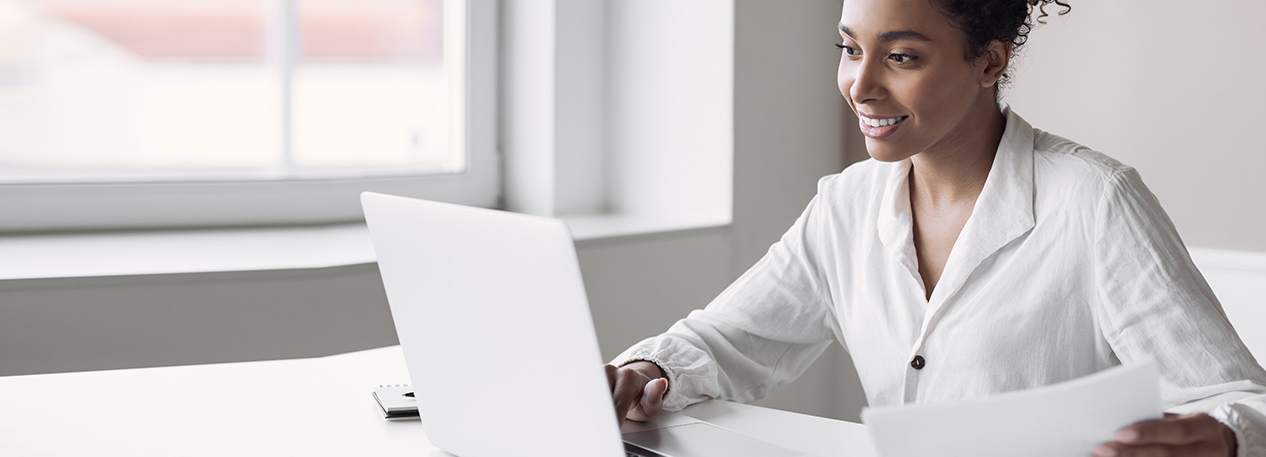 A business woman dressed in white works on a laptop in front of a window.