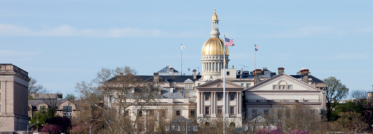 The American flag waves in the wind in front of the golden dome of the NJ state capital building.