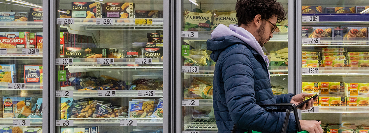 A man checks his cell phone while browsing the freezer section of the grocery store.