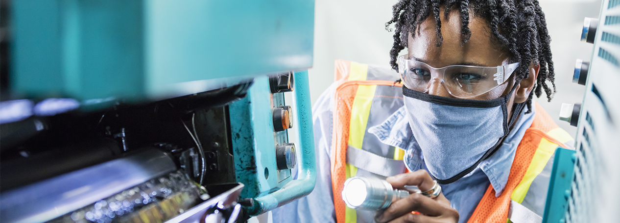 Woman in goggles and a face mask holding a flashlight and inspecting a machine.