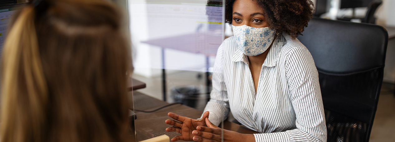 A masked woman leads a conversation with another across a desk. They are separated by a plastic partition.