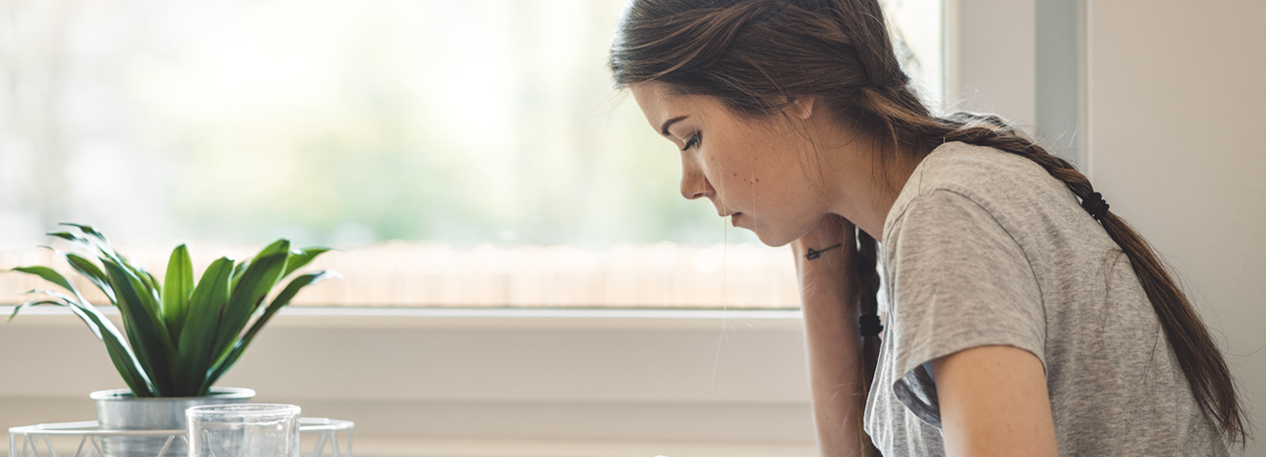 A young woman sits at a kitchen table next to a window and reads information on her cell phone.