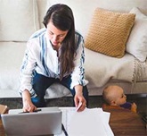 Woman sitting on a couch working on a laptop and papers with a small child next to her