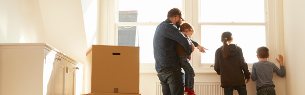 Man and three children looking out of a window in a room with moving boxes stacked on the floor.