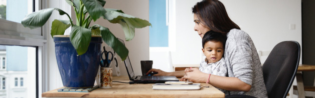 Woman working at home, looking at a piece of paper, holding a young child on her lap.