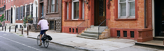 A man rides a bike down a city street along a block of brick townhouses.